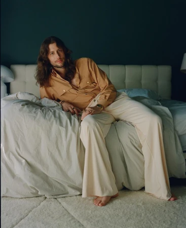 A fair-skinned man with long brown hair in an orange shirt and cream pants reclines on a bed with a blue wall in the background.