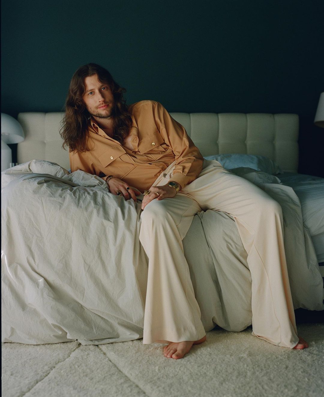 Portrait of a brunette male model with long hair, wearing a tan button-down shirt and cream-colored pants, posing on a bed indoors.