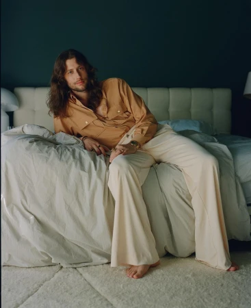 Portrait of a brunette male model with long hair, wearing a tan button-down shirt and cream-colored pants, posing on a bed indoors.