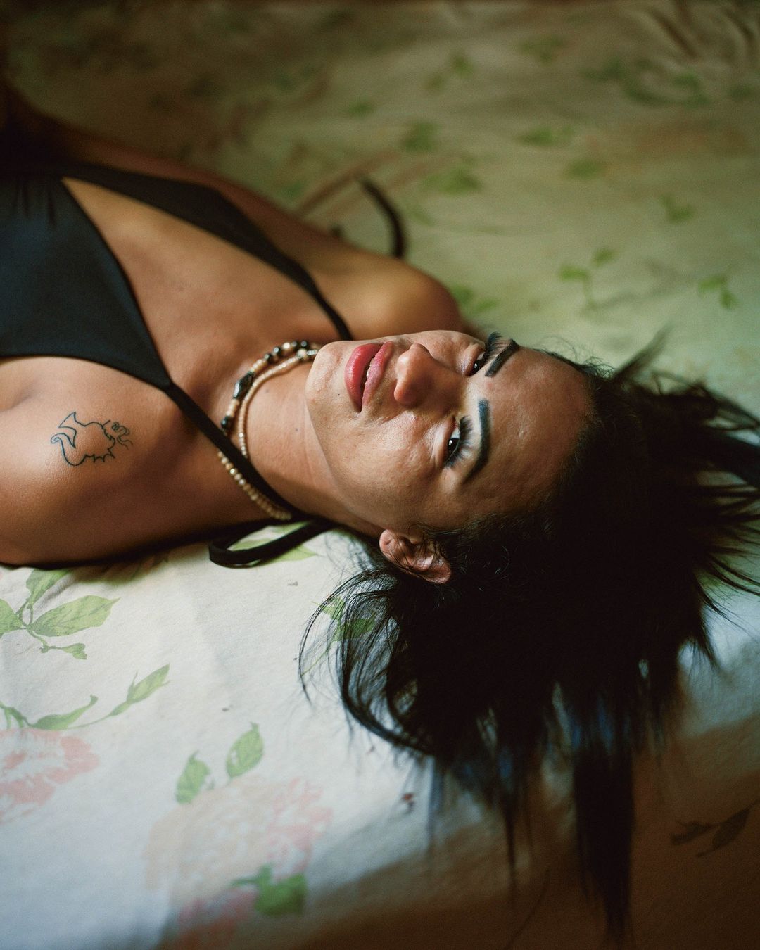 Close-up portrait of a woman lying on a patterned bed wearing a black top and beaded necklaces.