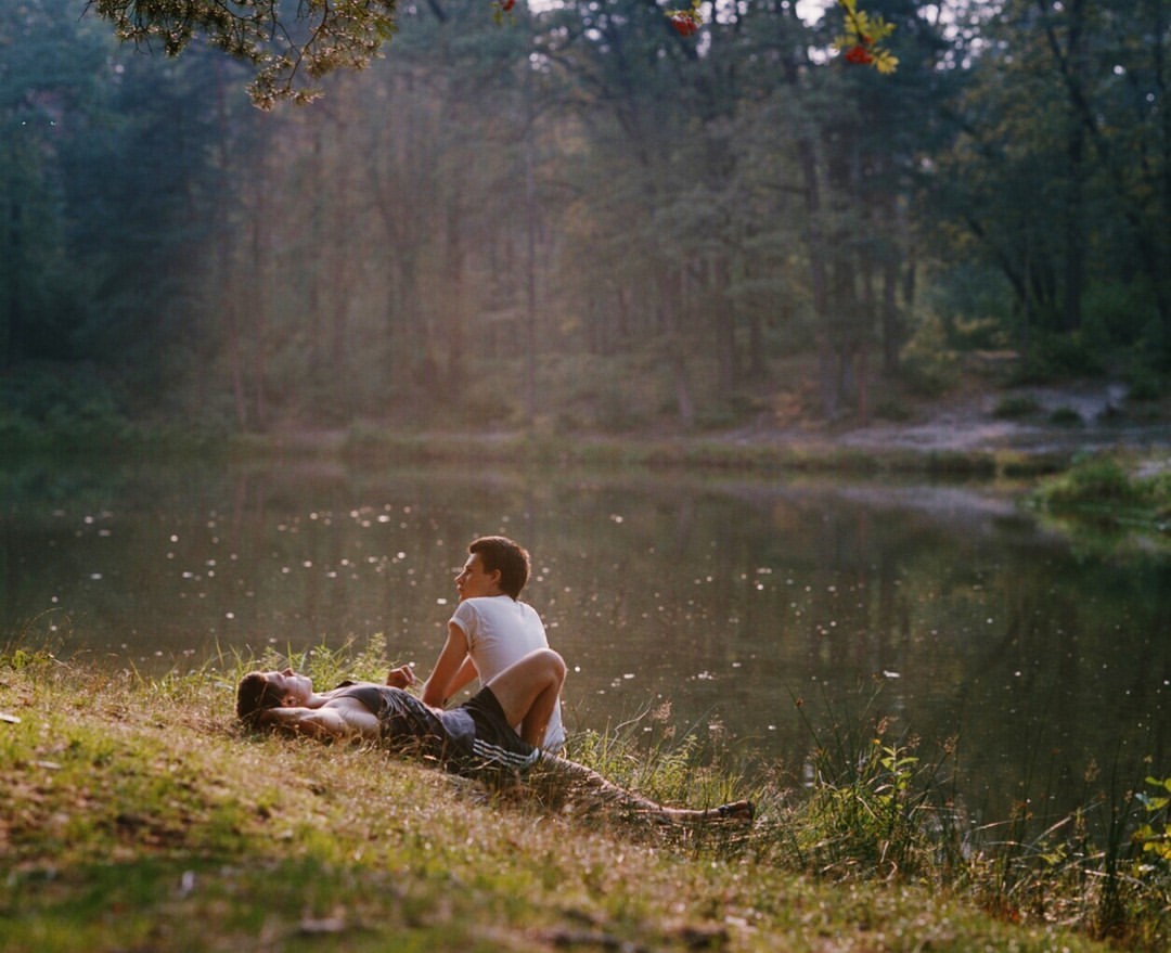 Two young adults relax on a grassy bank near a serene lake surrounded by a lush green forest in soft, natural light, creating a peaceful scene.