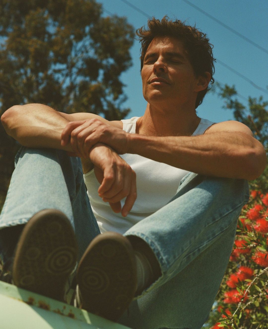 James Marsden in a white tank top and blue jeans sitting outdoors with eyes closed under a clear blue sky
