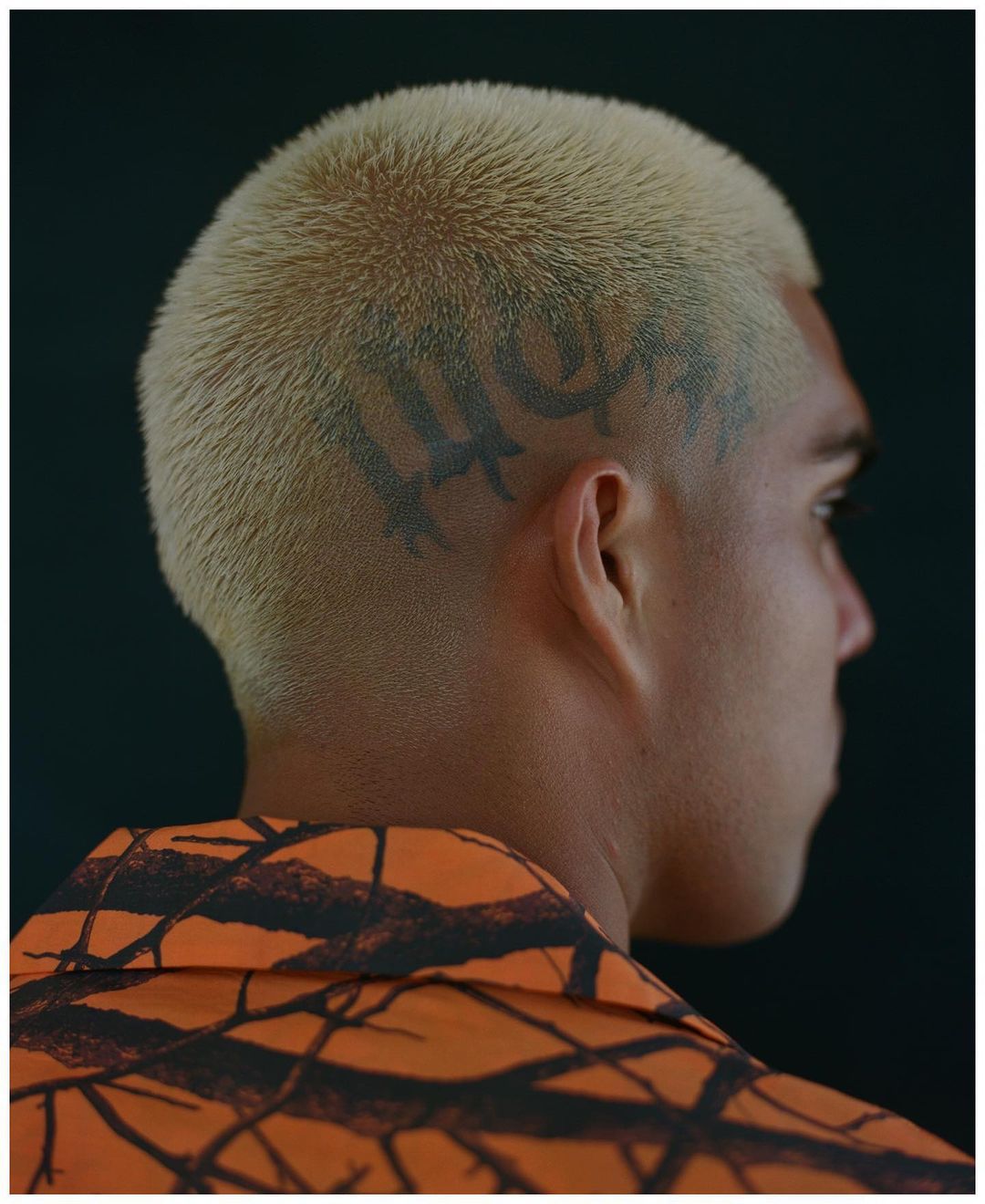 Profile of adult male with blonde hair and tattoo, wearing an orange patterned shirt, against a dark background in eye-level shot.