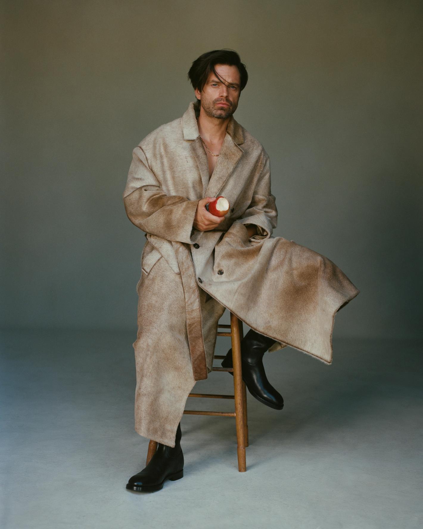 A fair-skinned man in a weathered coat seated on a wooden stool, holding an apple in a photo studio against a neutral background.