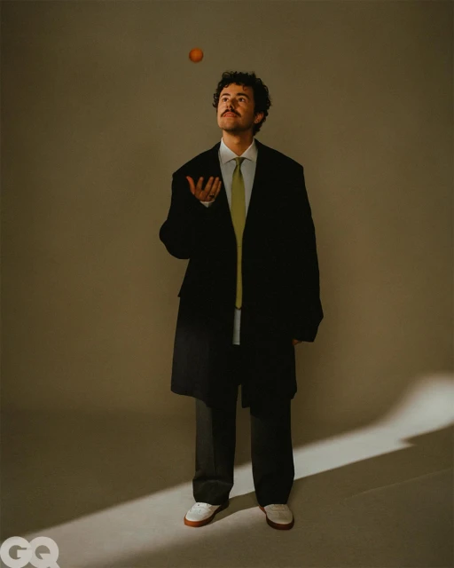Portrait of a man juggling an orange in a studio. He's dressed in a black jacket and tie, looking upwards with a neutral expression.