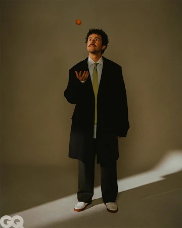 Portrait of a man juggling an orange in a studio. He's dressed in a black jacket and tie, looking upwards with a neutral expression.