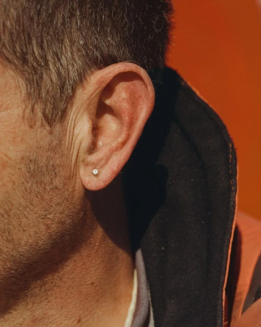 Close-up of a man's ear with a small earring, set against a bright orange backdrop, shows detail of skin texture and short hair.