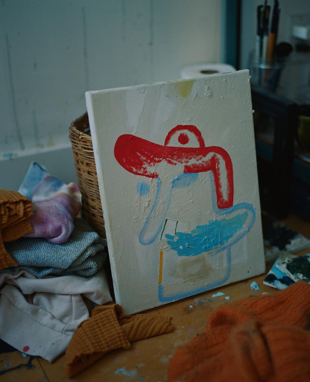 Abstract painting on canvas on a wooden floor, featuring red and blue shapes, surrounded by folded textile items and painting tools in a room.