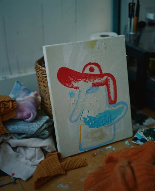 Abstract painting on canvas on a wooden floor, featuring red and blue shapes, surrounded by folded textile items and painting tools in a room.