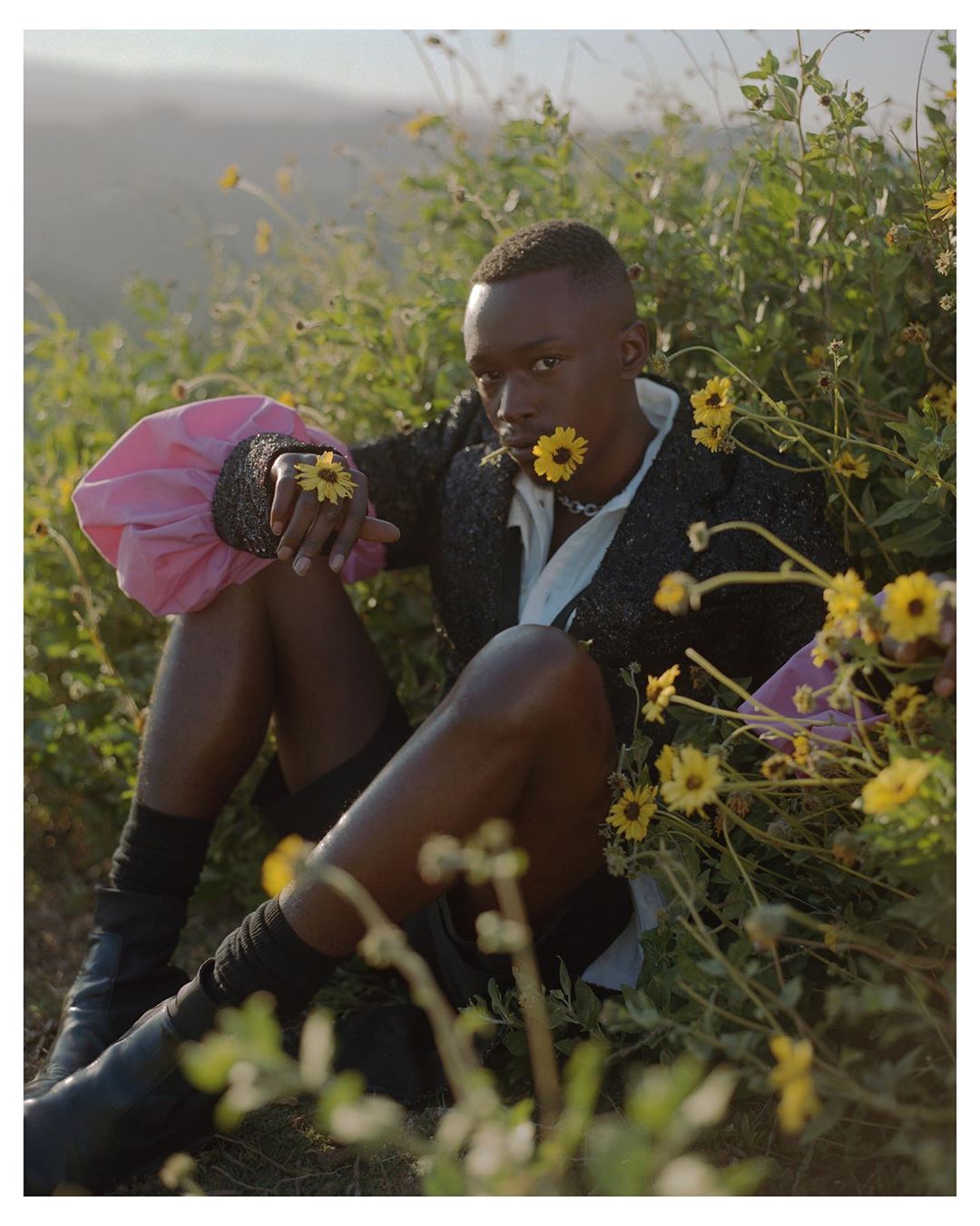 Portrait of a young Black man sitting in a field of yellow flowers wearing black boots, shorts, and a pink-sleeved jacket.