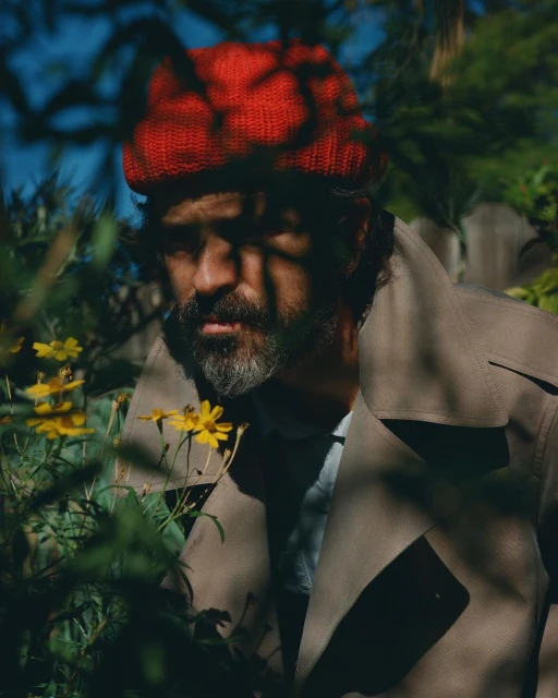 Man in a red beanie and trench coat stands among green plants and yellow flowers in an outdoor garden setting with natural light.