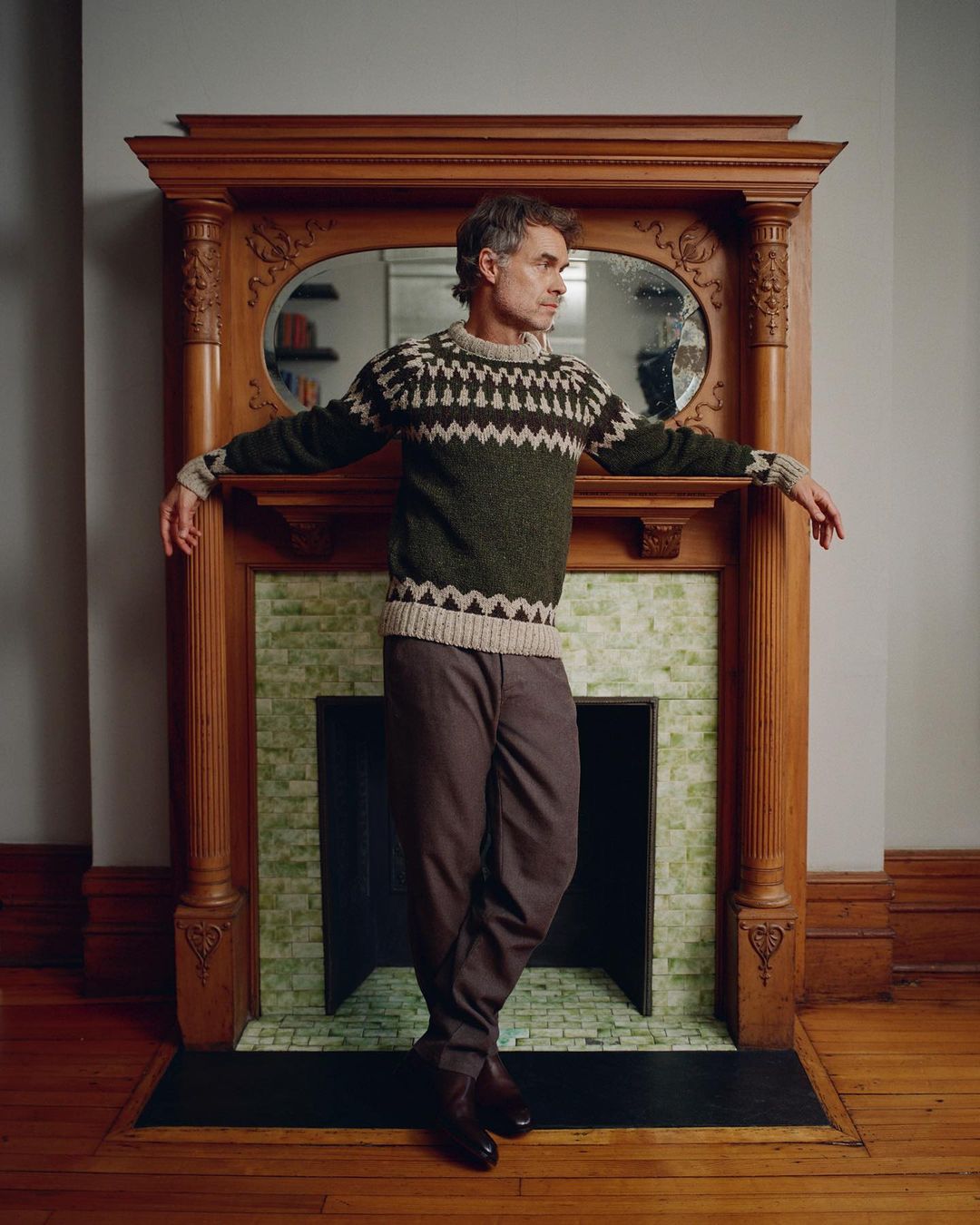Man in patterned sweater poses in front of decorative fireplace with wood mantel in an indoor setting, looking off to the side.