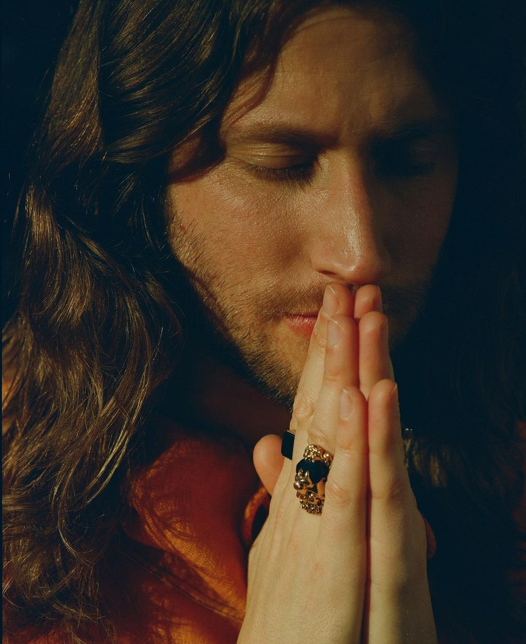 Portrait of a man with long brown hair, eyes closed, and hands together in prayer, featuring prominent gold rings against a dark background.