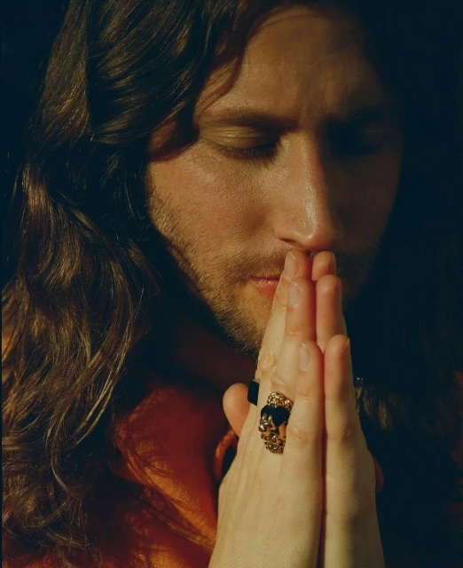 Portrait of a man with long brown hair, eyes closed, and hands together in prayer, featuring prominent gold rings against a dark background.