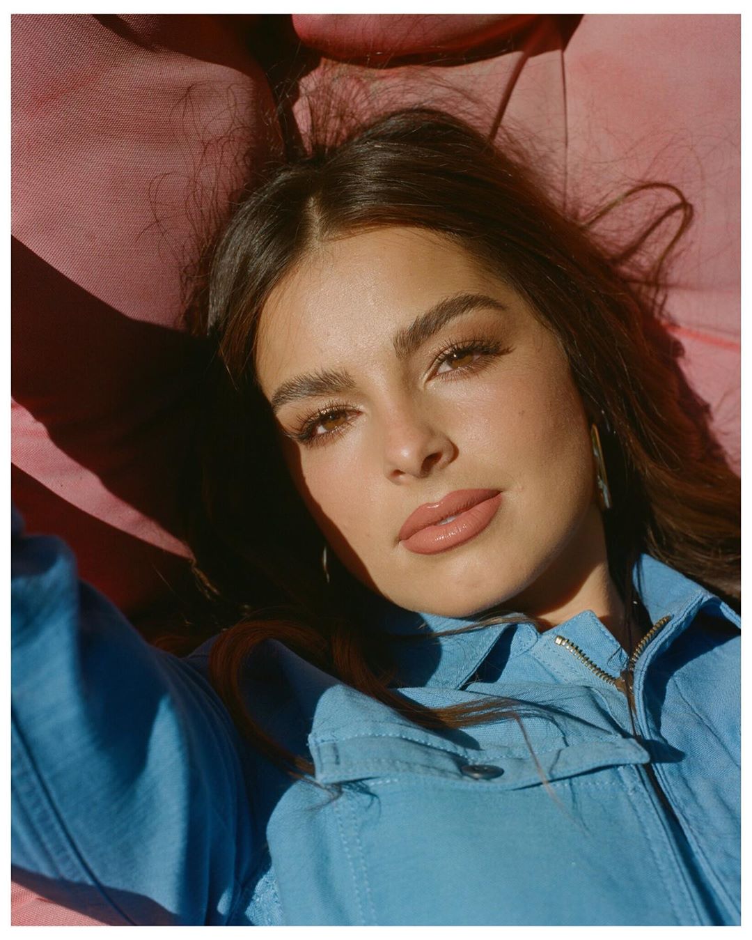 Close-up portrait of a woman with light brown hair and blue denim jacket, lying on a pink fabric surface in soft, diffused lighting.