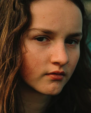 Close-up portrait of a young person with long brown hair, freckles, and a serious expression captured by Daniel Jack Lyons.