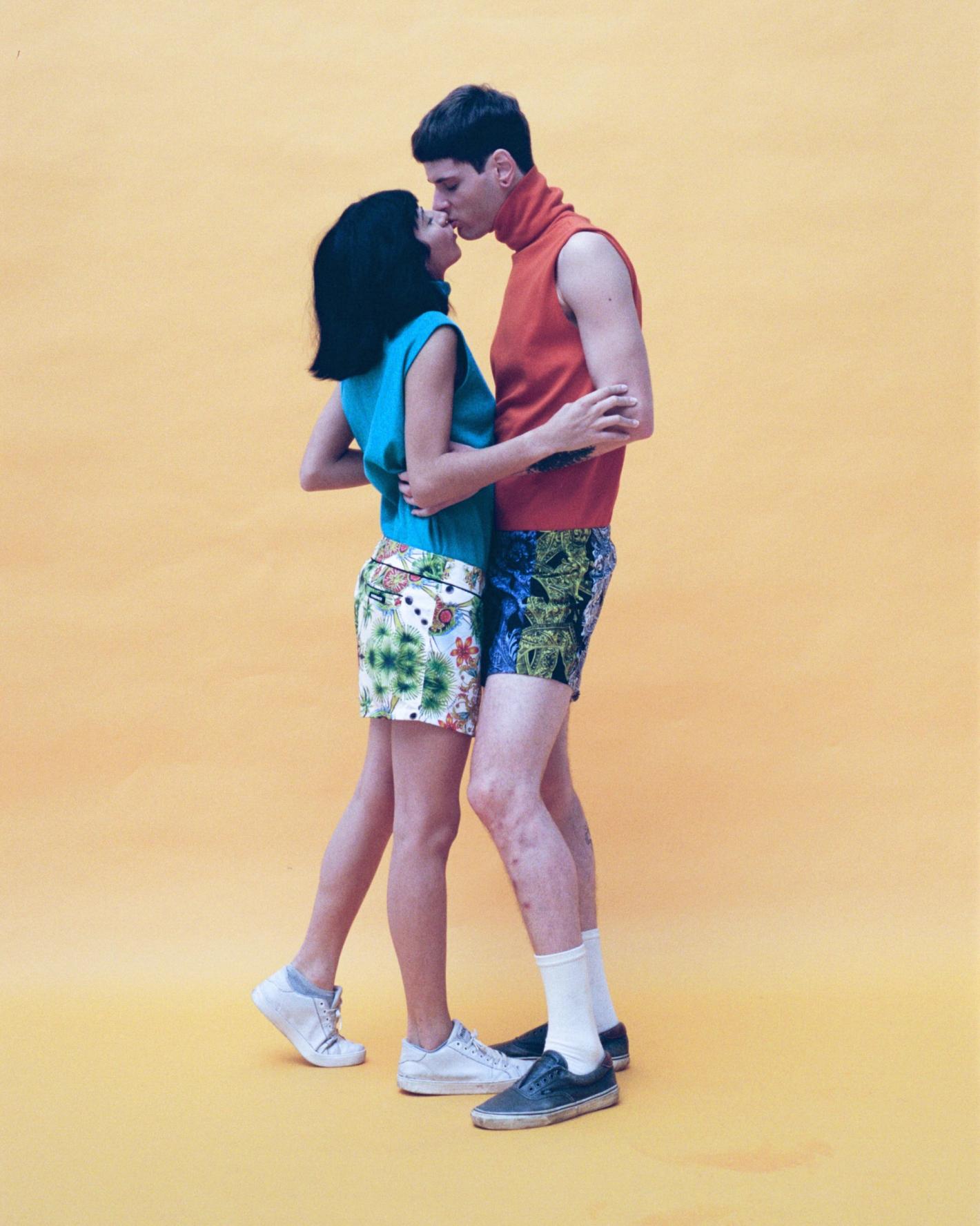 Man and woman standing and kissing against a solid yellow background wearing colorful casual clothing