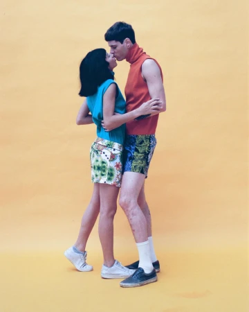 Man and woman standing and kissing against a solid yellow background wearing colorful casual clothing