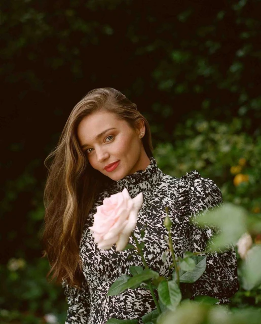 Portrait of a woman with long brown hair in a black-and-white dress, holding a light pink rose in an outdoor garden setting.