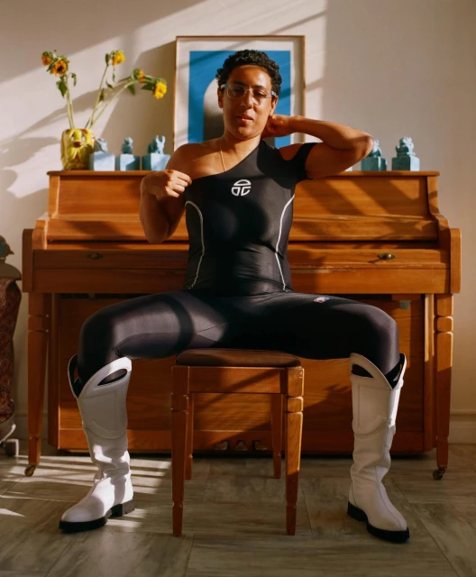 Woman in athletic wear sits on wooden stool in front of piano with sunflowers and framed art in background in sunlit room.