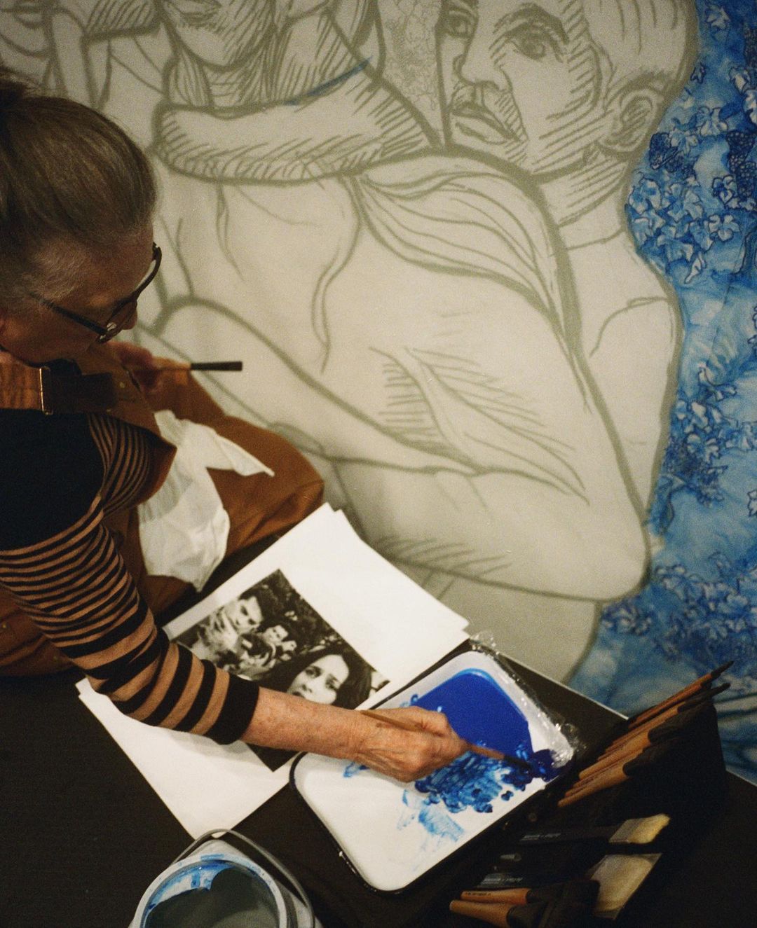 Artist at work on a large mural, painting a sketch of figures with blue paint; indoors, daytime, art technique, artist, mural.