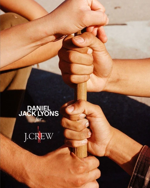 Several hands of different skin tones hold a wooden stick, with a logo of 'DANIEL JACK LYONS for J.CREW' visible in the frame.