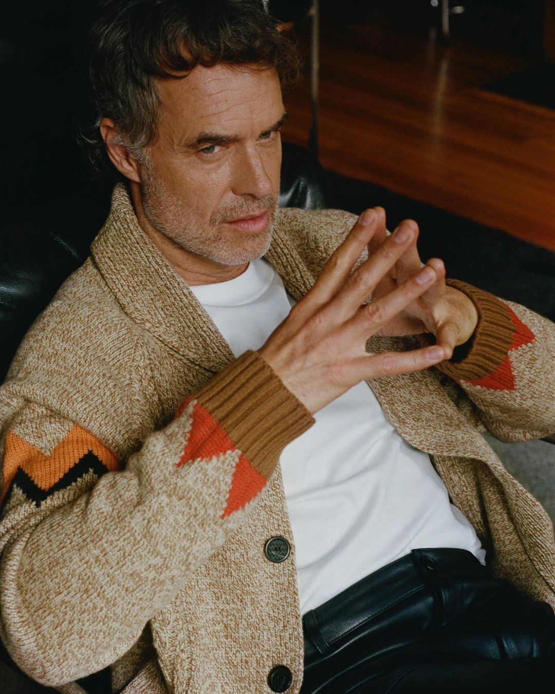 Portrait of an adult male sitting with hands clasped, wearing an open knit cardigan sweater, white t-shirt, and black pants in an indoor setting.