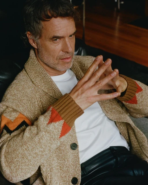 Portrait of an adult male sitting with hands clasped, wearing an open knit cardigan sweater, white t-shirt, and black pants in an indoor setting.