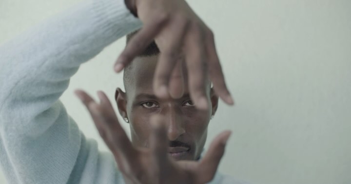 Close-up portrait of a dark-skinned adult male framing his face with his fingers indoors against a neutral background, wearing a blue sweater.