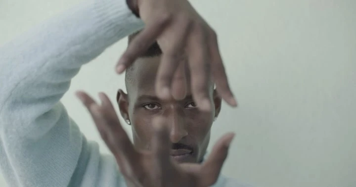 Close-up portrait of a dark-skinned adult male framing his face with his fingers indoors against a neutral background, wearing a blue sweater.