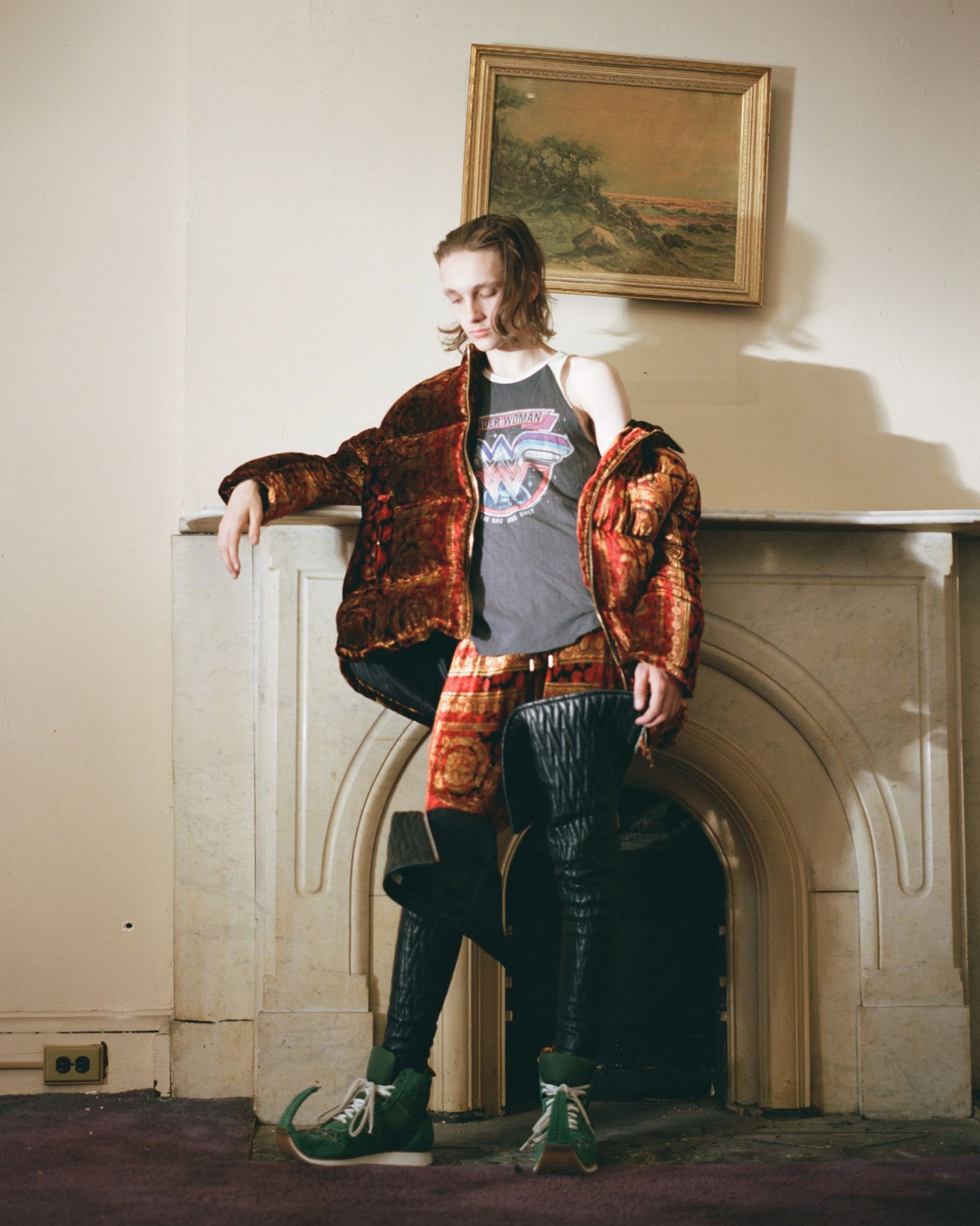 A person wearing high fashion leans against a white fireplace indoors with an art piece displayed above the mantel.