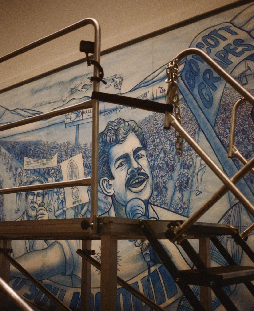 Illustration of Cesar Chavez speaking at a rally with supporters holding signs, depicted in blue and white on a mural.