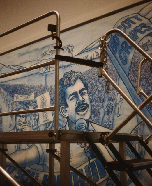 Illustration of Cesar Chavez speaking at a rally with supporters holding signs, depicted in blue and white on a mural.