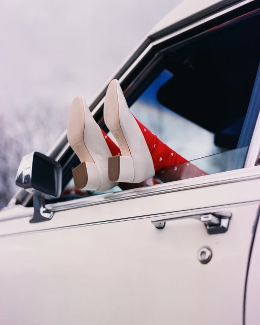Legs wearing white heels and red patterned tights are propped out of the car window. The vintage car is white and set against a pale sky.