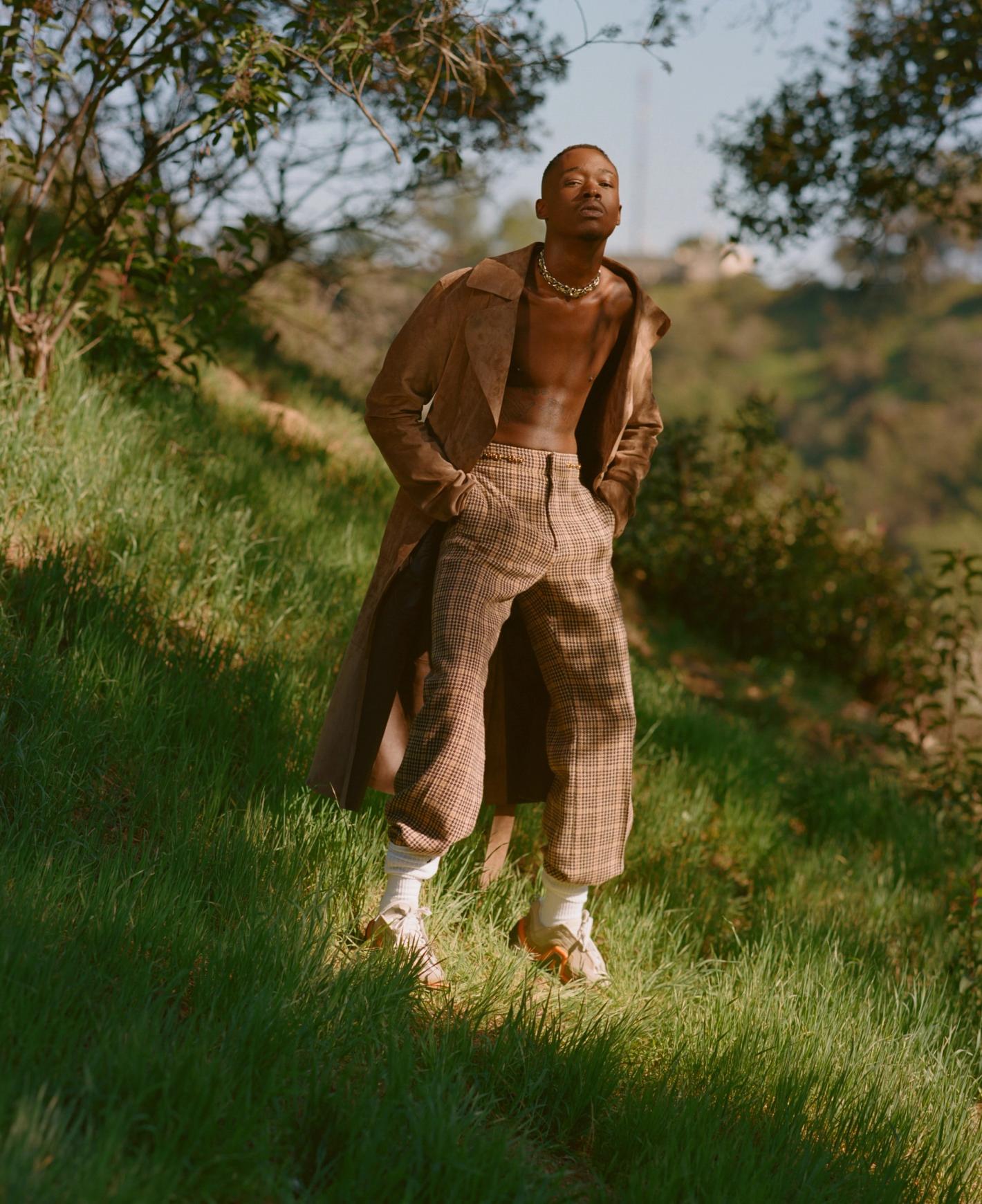 Stylish man poses on grassy hill wearing a brown suede coat and patterned pants, complemented by socks and sandals in a natural setting.