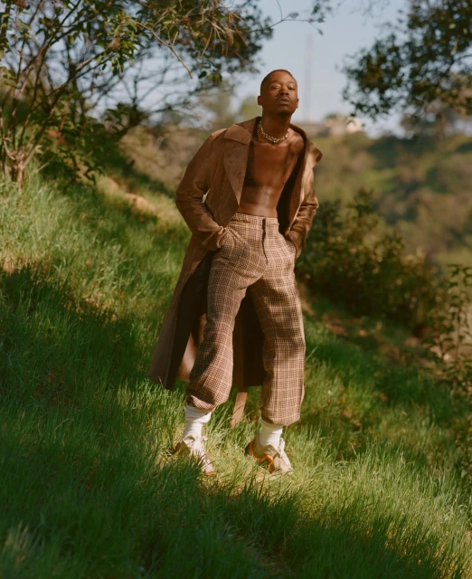 Stylish man poses on grassy hill wearing a brown suede coat and patterned pants, complemented by socks and sandals in a natural setting.