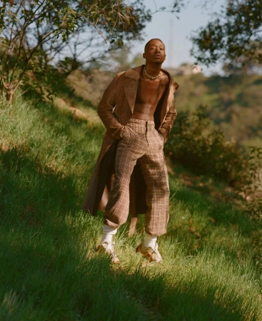 Stylish man poses on grassy hill wearing a brown suede coat and patterned pants, complemented by socks and sandals in a natural setting.