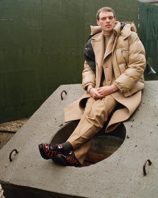 Man in beige puffer jacket and overcoat sits on concrete structure outdoors with textured green background, wearing dark sneakers.