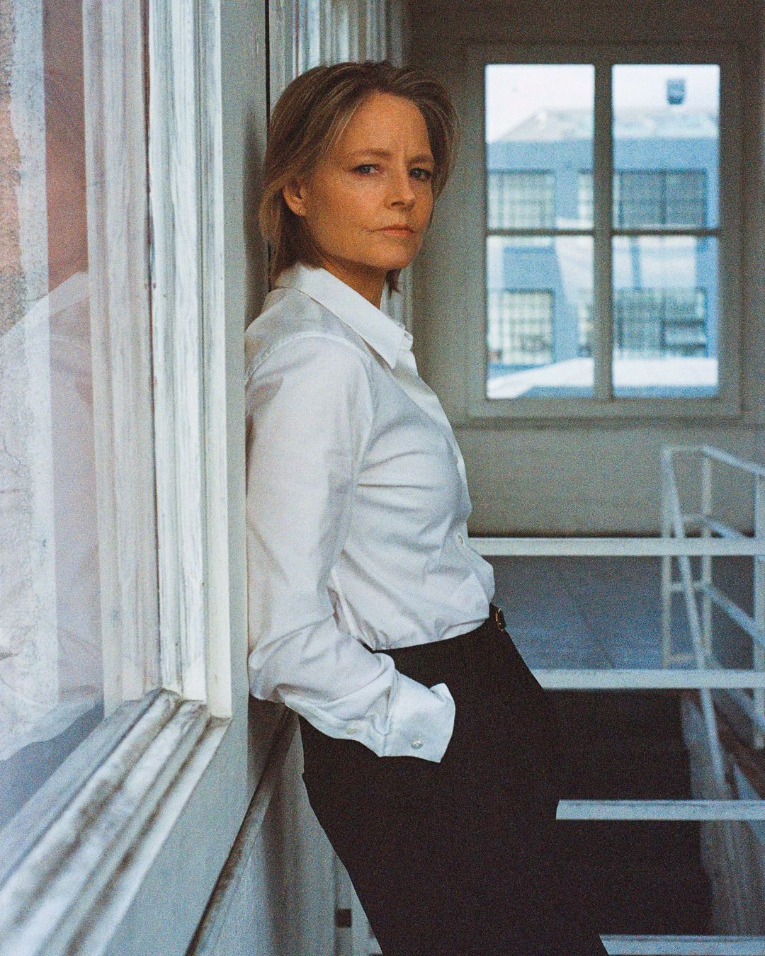 Headshot of adult woman in a white blouse and black slacks leaning against window frame with building visible through a window in background.