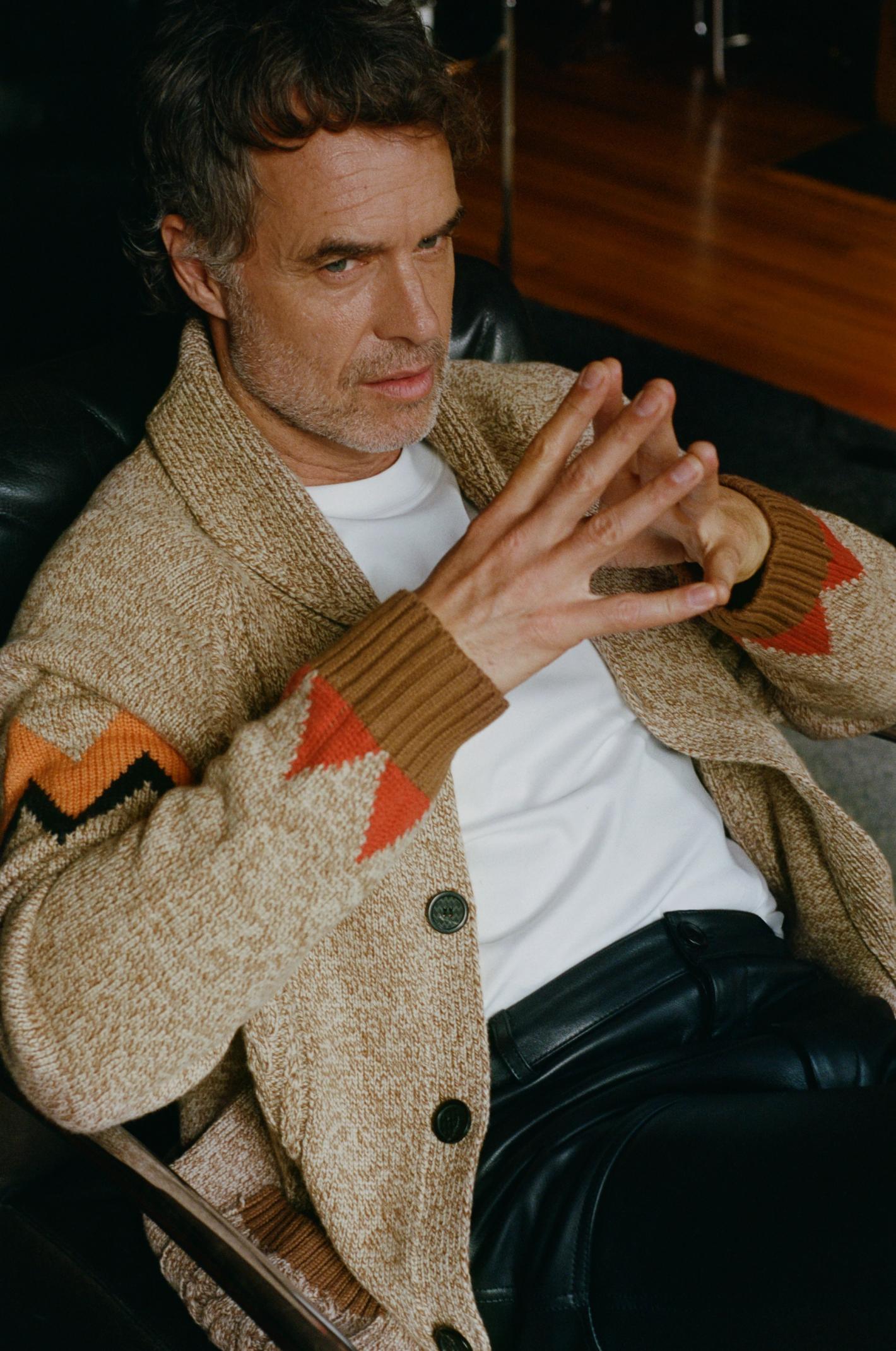 Adult male in a white t-shirt and patterned cardigan poses while seated indoors, his fingers interlaced, against a blurred background.