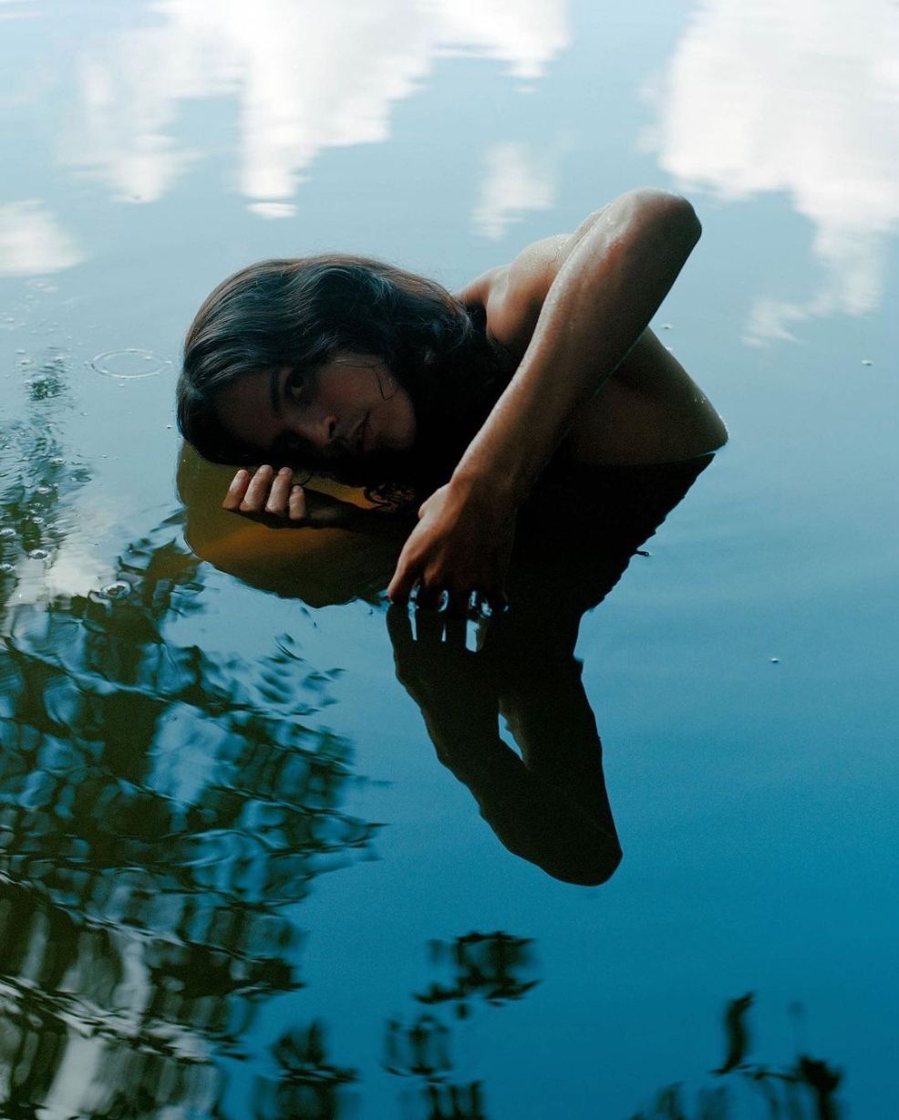 Adult with long dark hair rests in calm water, reflection visible, against background of sky reflections and trees in tranquil outdoor scene.