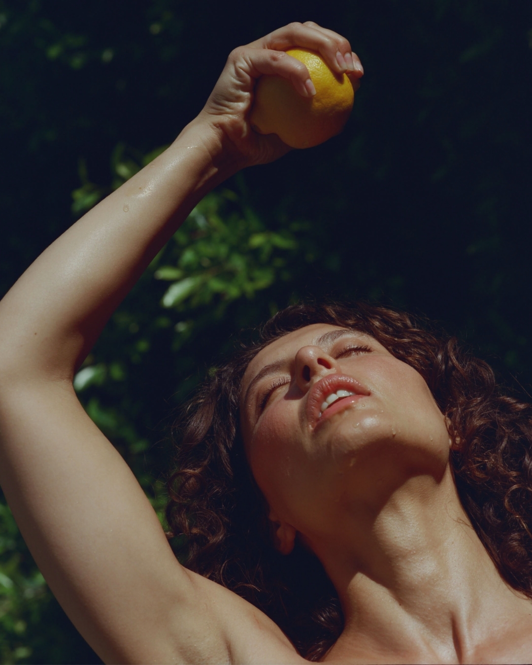 Woman squeezes a lemon above her face, eyes closed and head tilted back. Sunlight glistens on her skin in an outdoor setting.