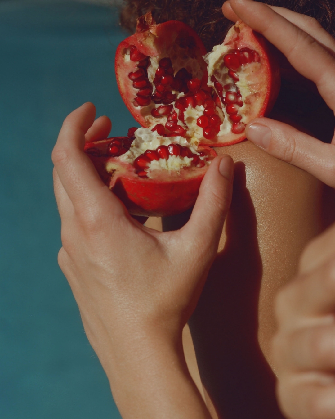 Close-up of a woman holding a halved pomegranate with red seeds, with a blurred blue background, highlighting the fruit's vibrant color.