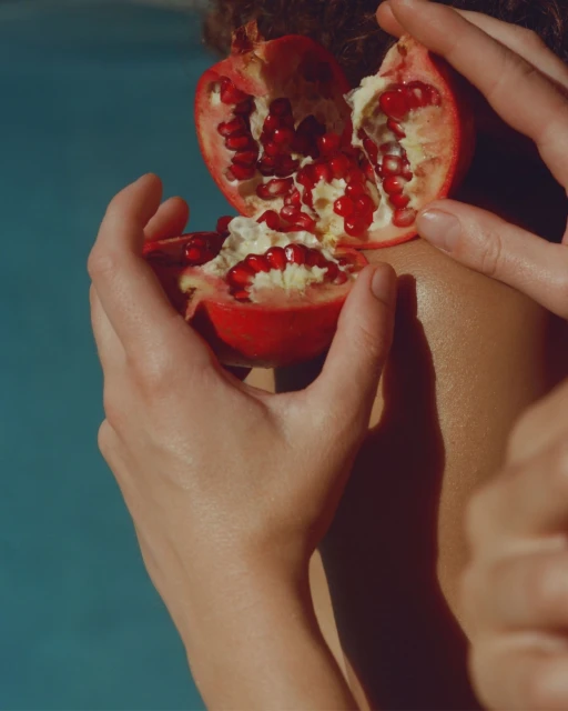 Close-up of a woman holding a halved pomegranate with red seeds, with a blurred blue background, highlighting the fruit's vibrant color.