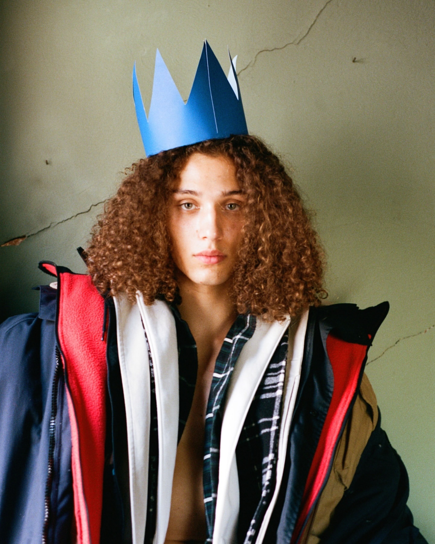 Portrait of a young adult man with curly hair wearing a blue paper crown and layered jackets against a cracked green wall.