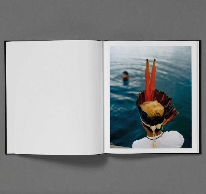 Open photo book showing a person wearing a traditional feathered headdress overlooking water with a swimmer