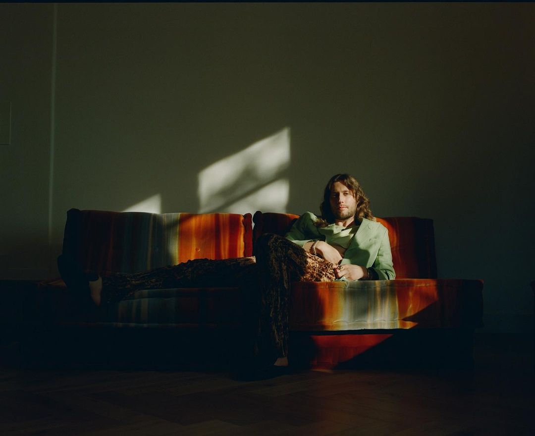 A man with long hair sits on a retro couch in a room with natural light, wearing a light green jacket and patterned pants.