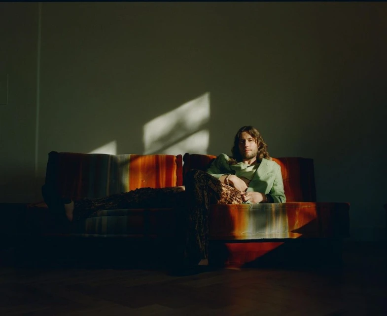 A man with long hair sits on a retro couch in a room with natural light, wearing a light green jacket and patterned pants.