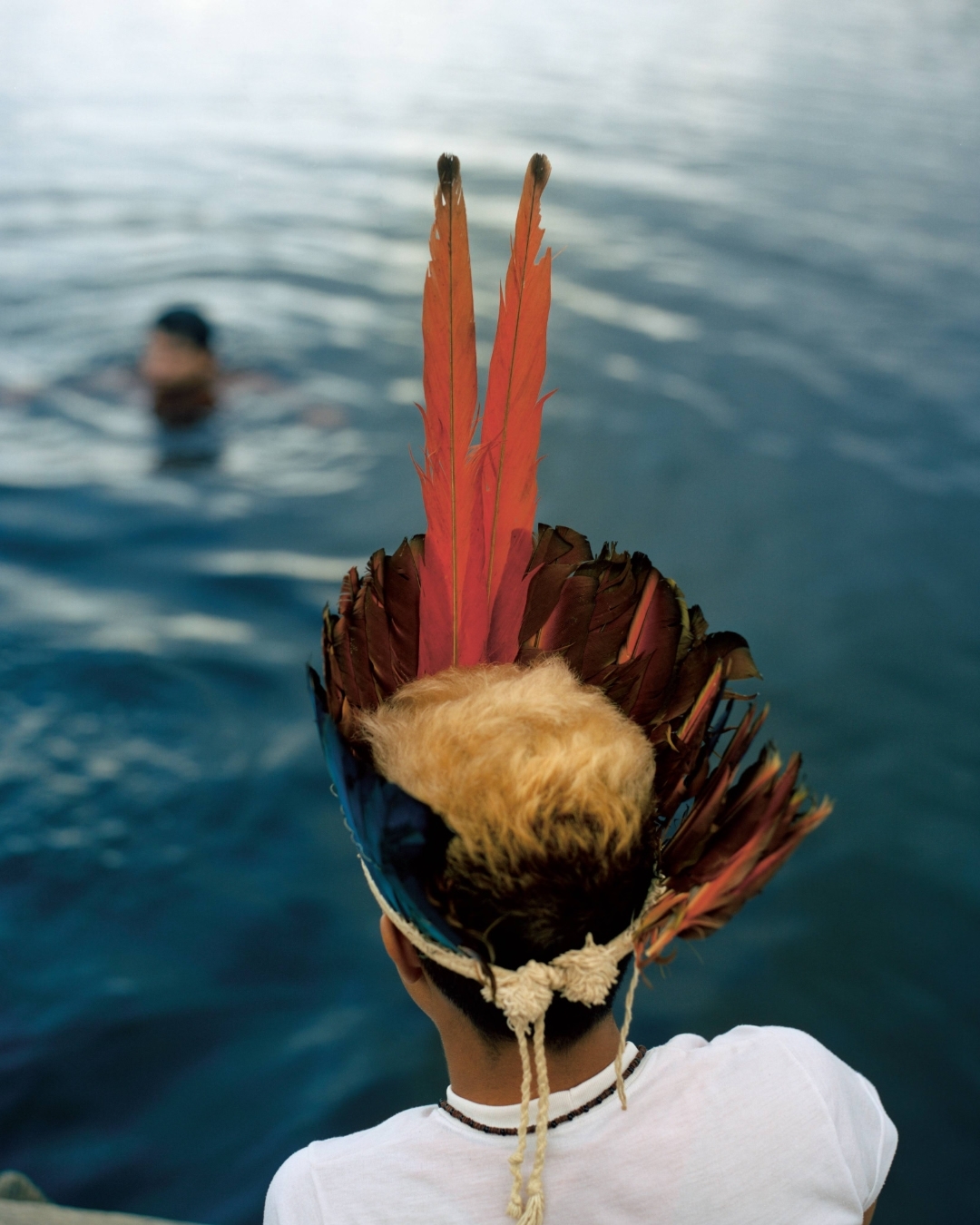 Indigenous youth with feather headdress viewed from behind near water, another person visible in background swimming in blue river.