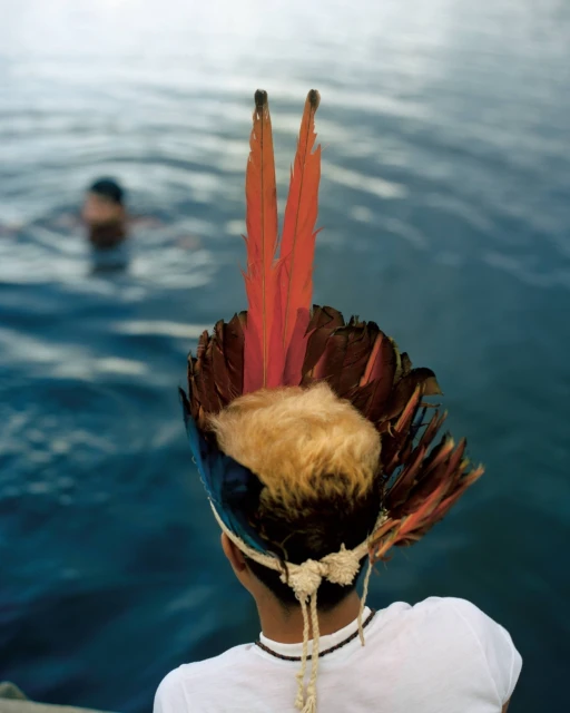 Indigenous youth with feather headdress viewed from behind near water, another person visible in background swimming in blue river.